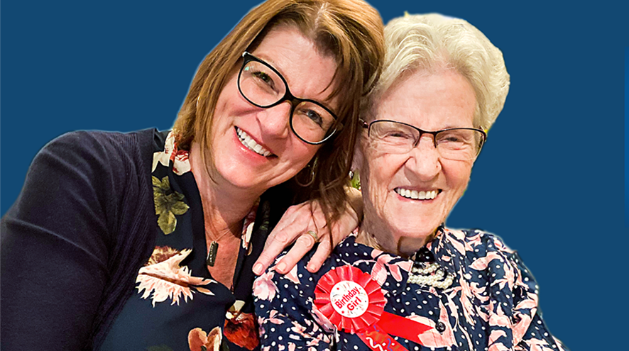 A middle-aged woman with long brown hair cuddles up with an older woman with a badge reading "Birthday girl" on her lapel.