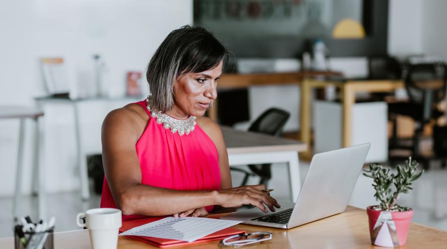Older woman with brown skin and greying dark hair, wearing a pink dress. She sits at a desk with an open laptop and paper files.